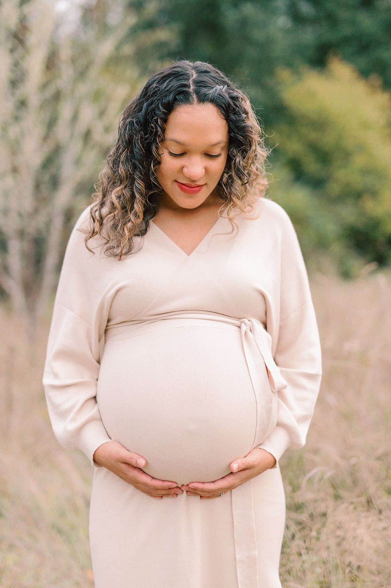 A mother to be in a cream maternity gown stands in a field of golden grass looking down at her bump Around the Circle Midwifery