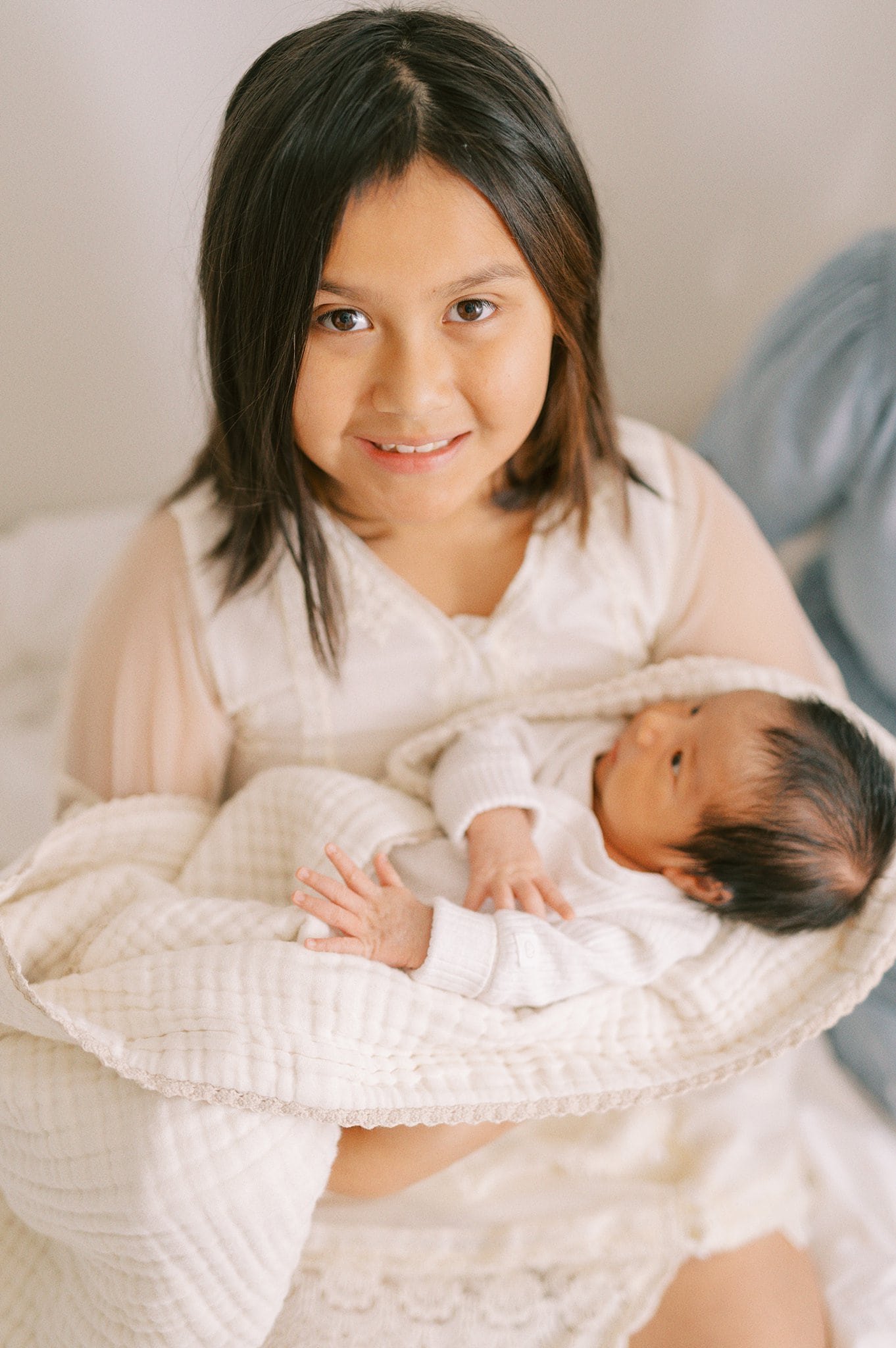 A young girl sits on a bed in a white lace dress while holding her newborn baby sibling Captain Little