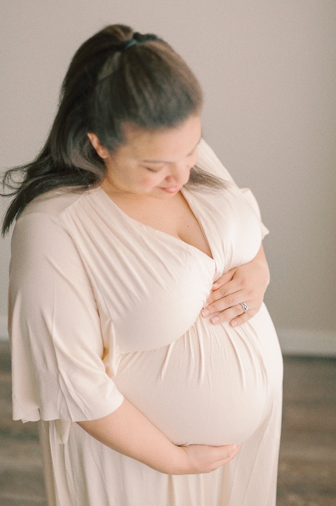 A mother to bee stands in a studio in a white maternity gown holding her bump