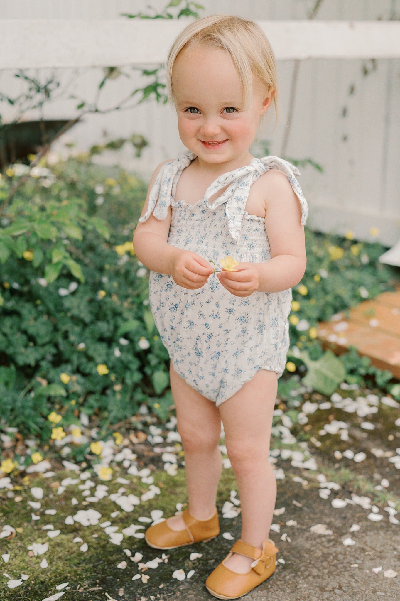 A young toddler in a onesie and leather shoes plays with flowers in a garden