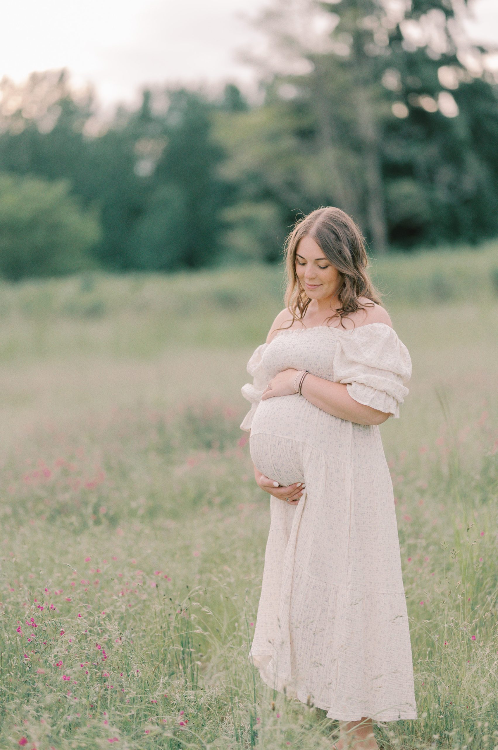 A pregnant woman in a white maternity dress stands in a field of wildflowers holding her bump Natural Beginnings Midwifery Care