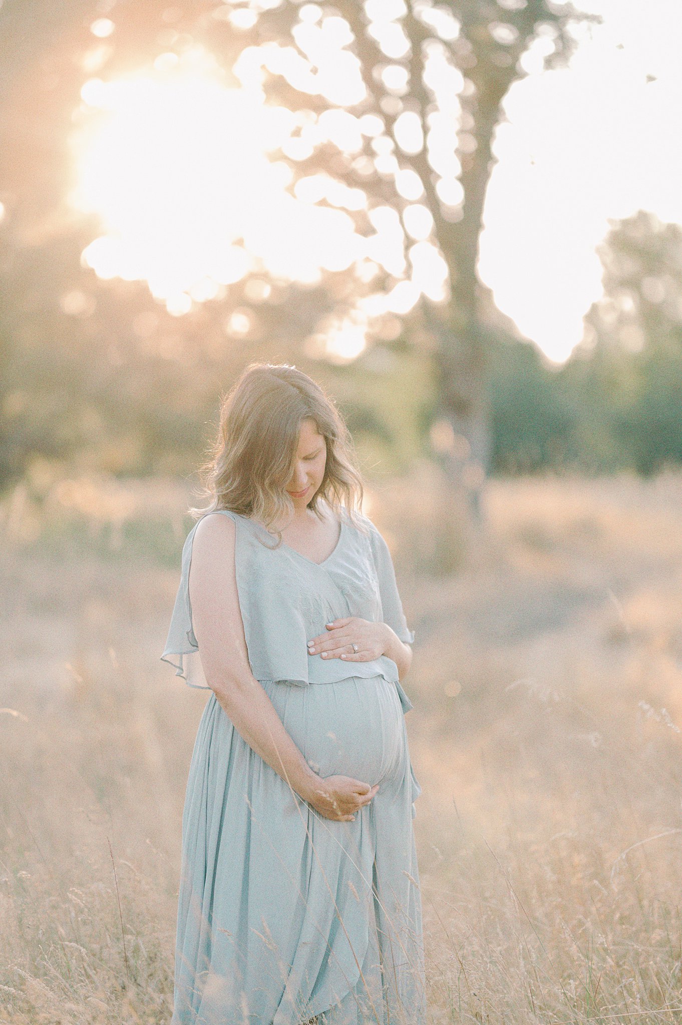 A mother to be gazes down at her bump in a blue maternity gown at sunset Tacoma Doulas