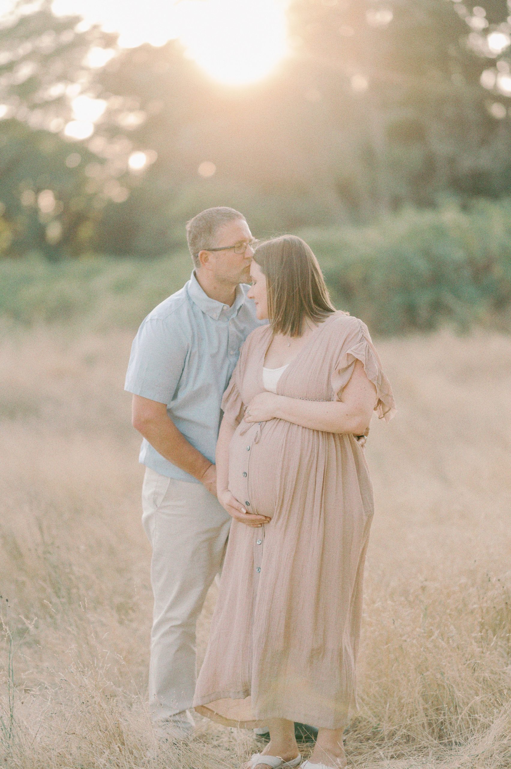 A mother to be in a beige dress holds her bump while her husband kisses her forehead in a field at sunset The Birthing Inn
