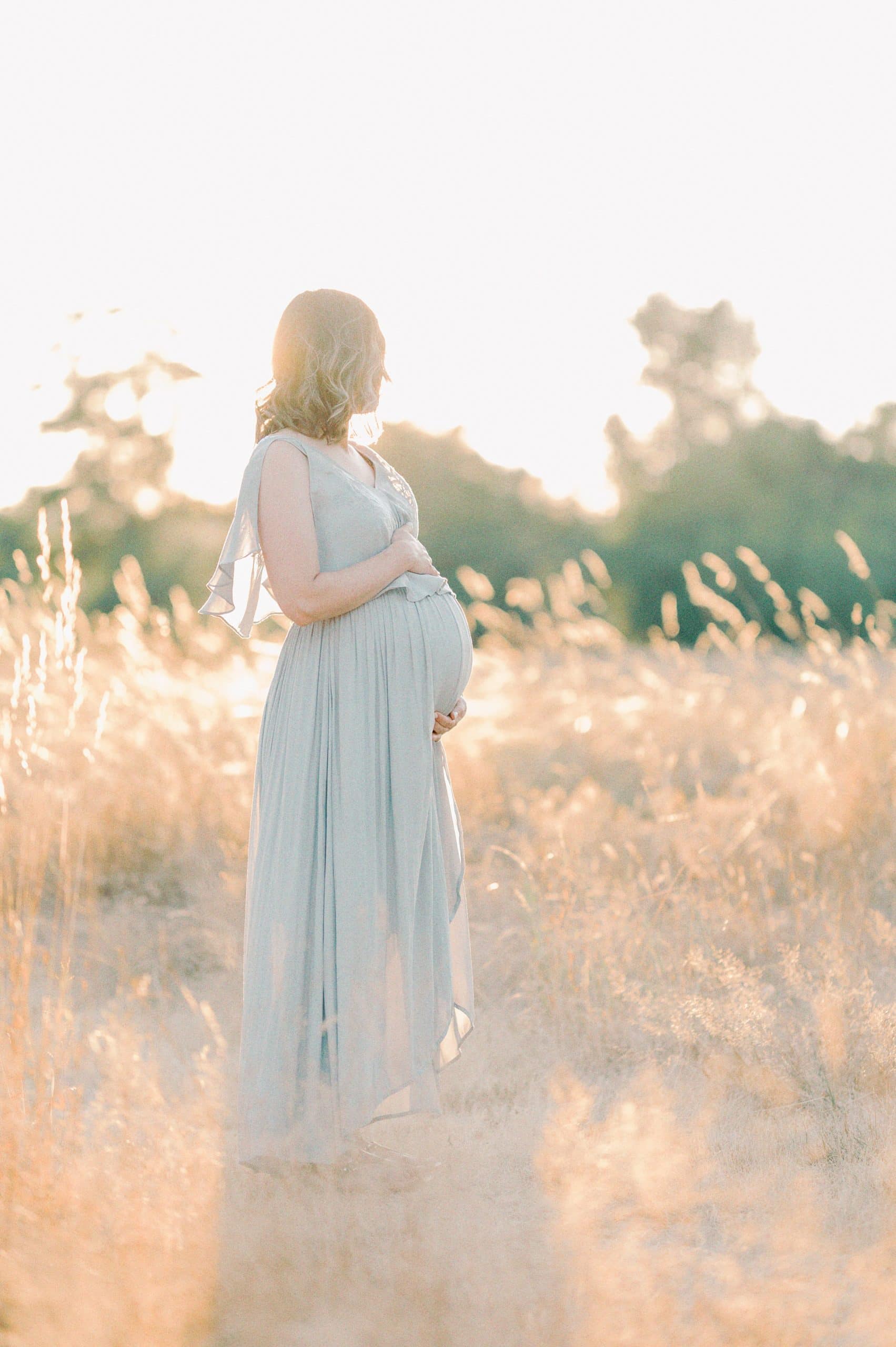 A mother to bee in a blue maternity gown stands in tall golden grass at sunset while looking off over her shoulder True Self Yoga Olympia