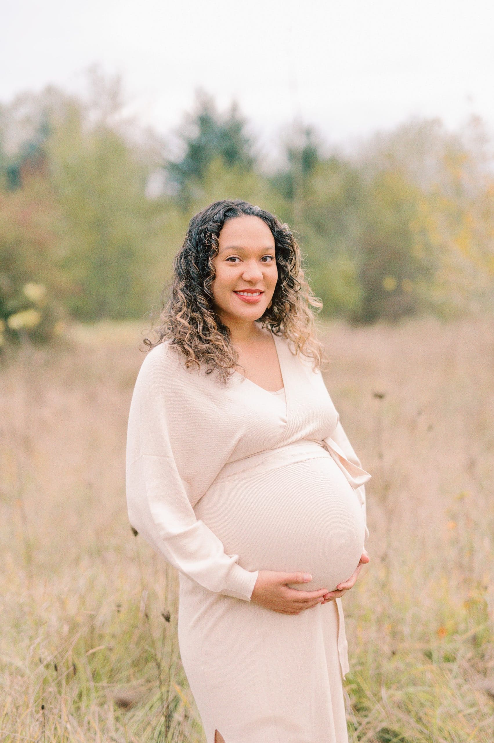 A mom to be stands in a field of tall grass while holding her bump in a beige maternity gown Womb revolution