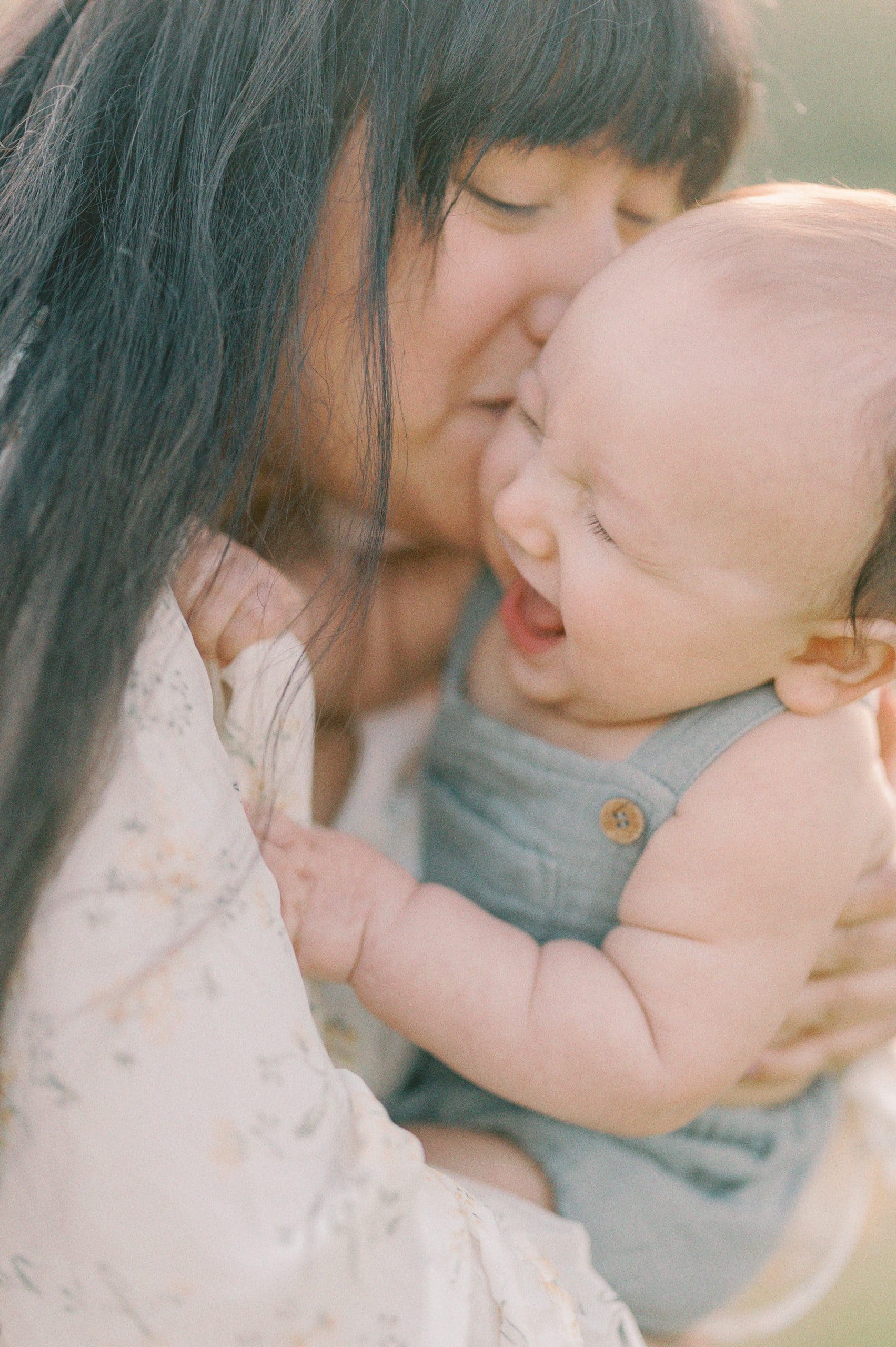 A mother kisses her young toddler baby outside wearing blue overalls Seattle Baby Boutiques