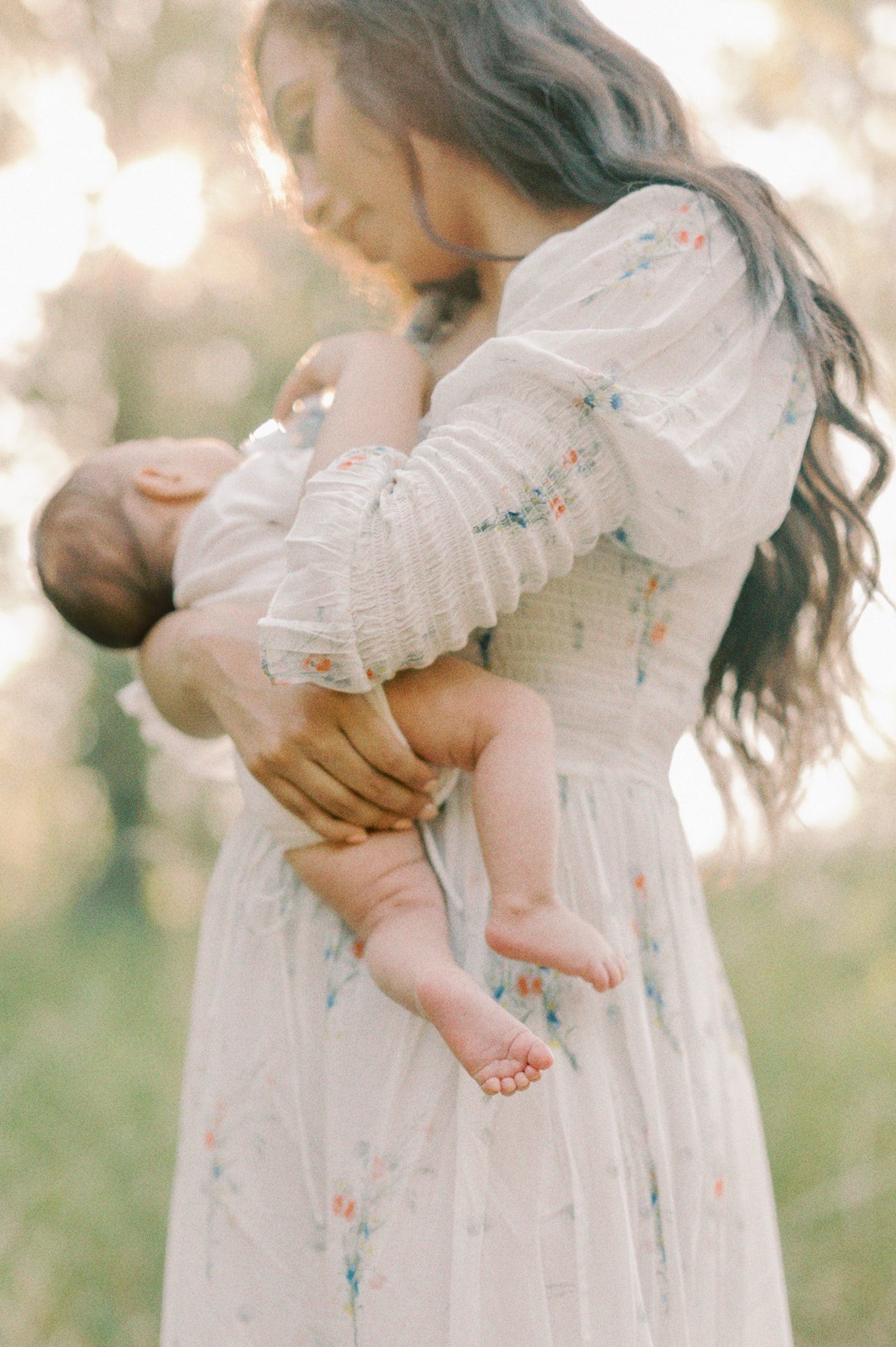 A mother stands in a forest holding her newborn baby in her arms in a white dress with florals Seattle Doulas