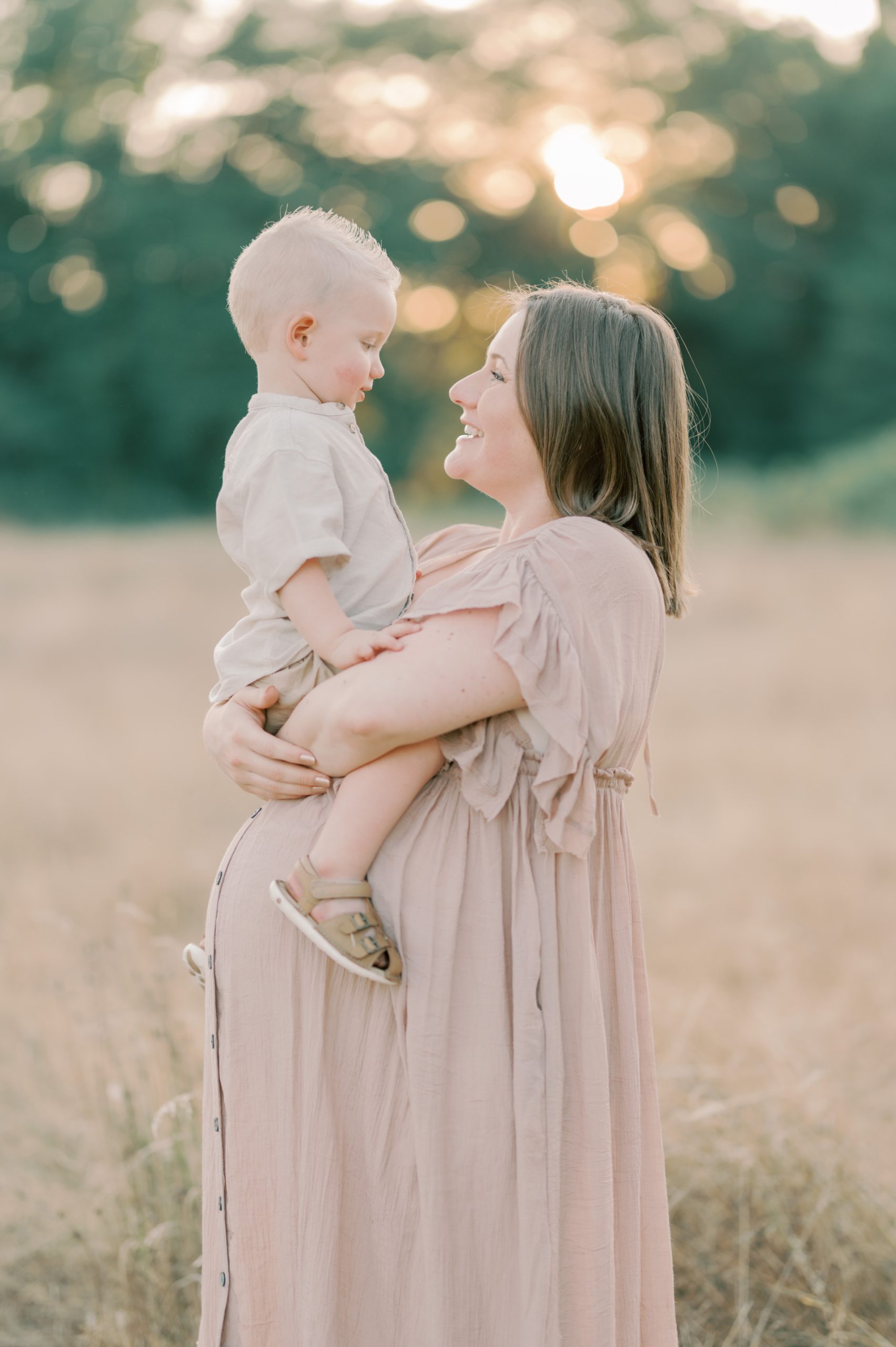 A pregnant mother stands in a field of tall golden grass with her toddler son sitting on her bump Seattle Placenta Encapsulation