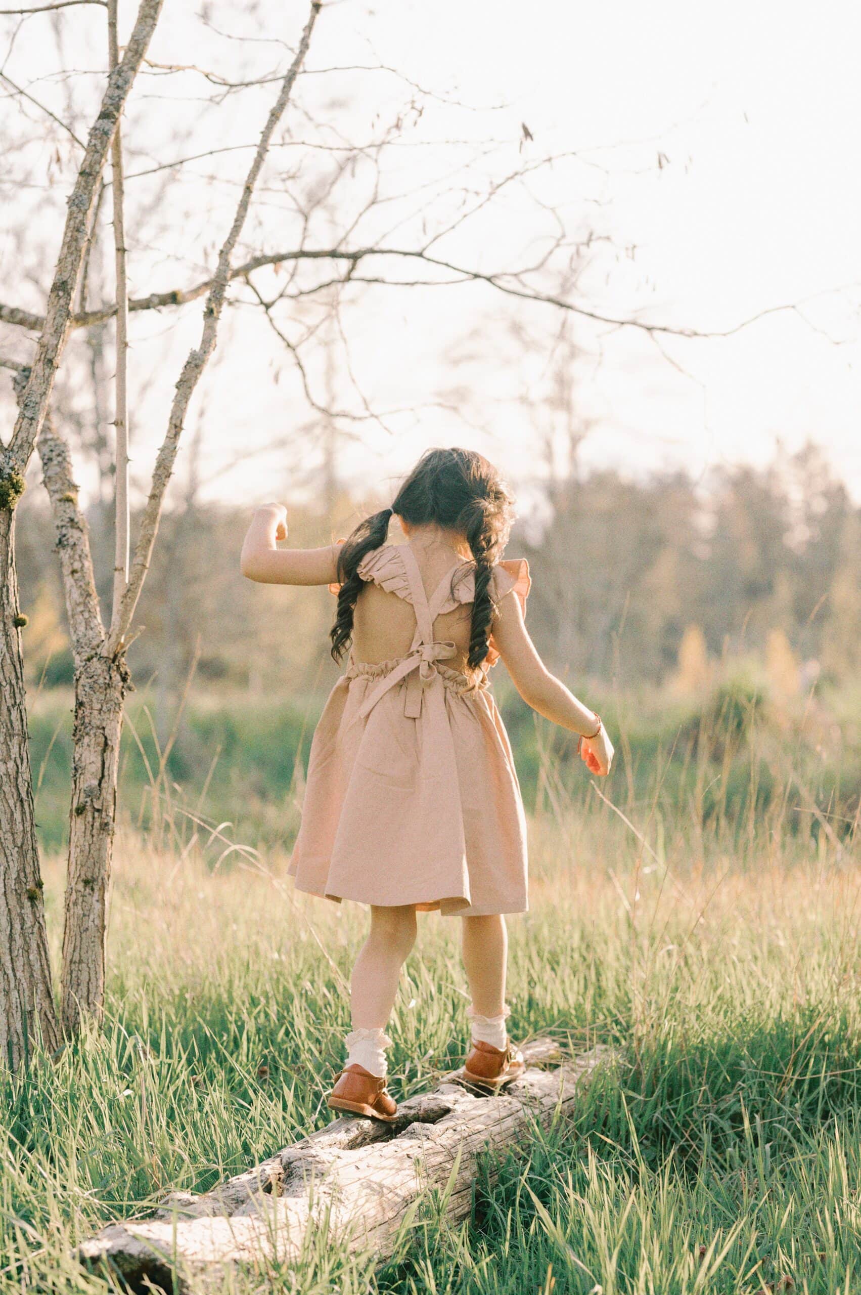 A young girl in a pink dress walks on a fallen log in a grassy park Seattle Preschools