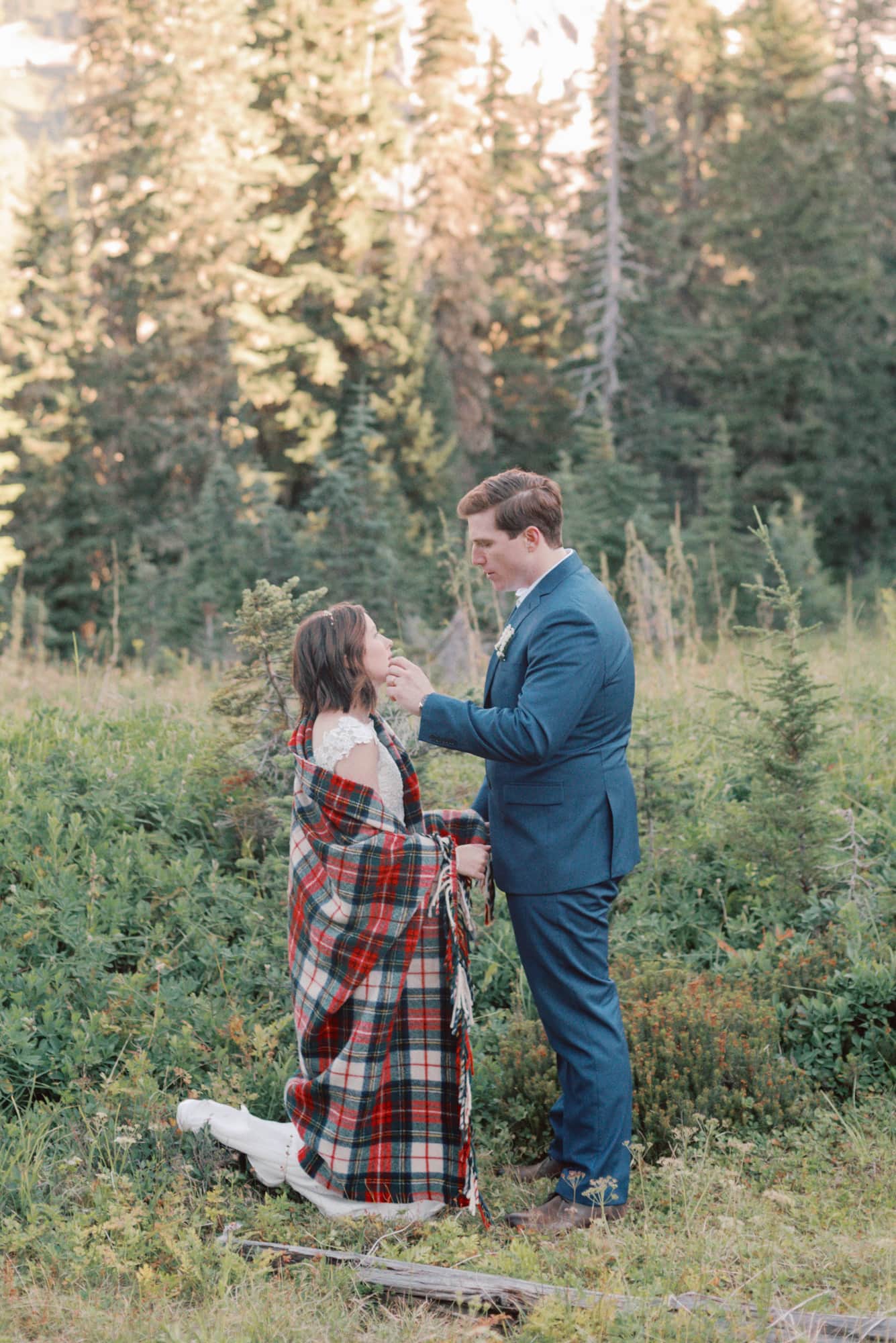 A bride wrapped in a red plaid blanket stands in a forest trail with her groom in a blue suit as he wipes something off her cheek