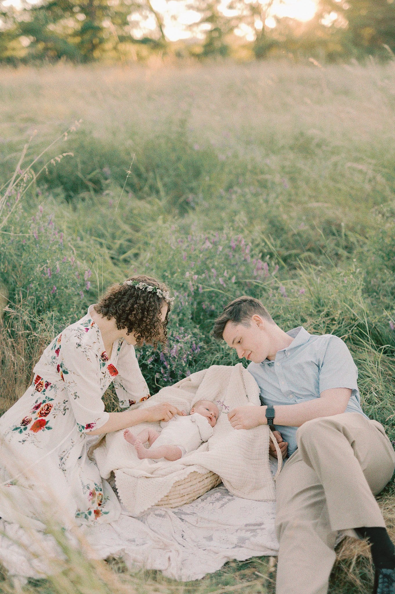 New parents lay on a picnic blanket with their newborn baby in a basket in a field of tall grass at sunset