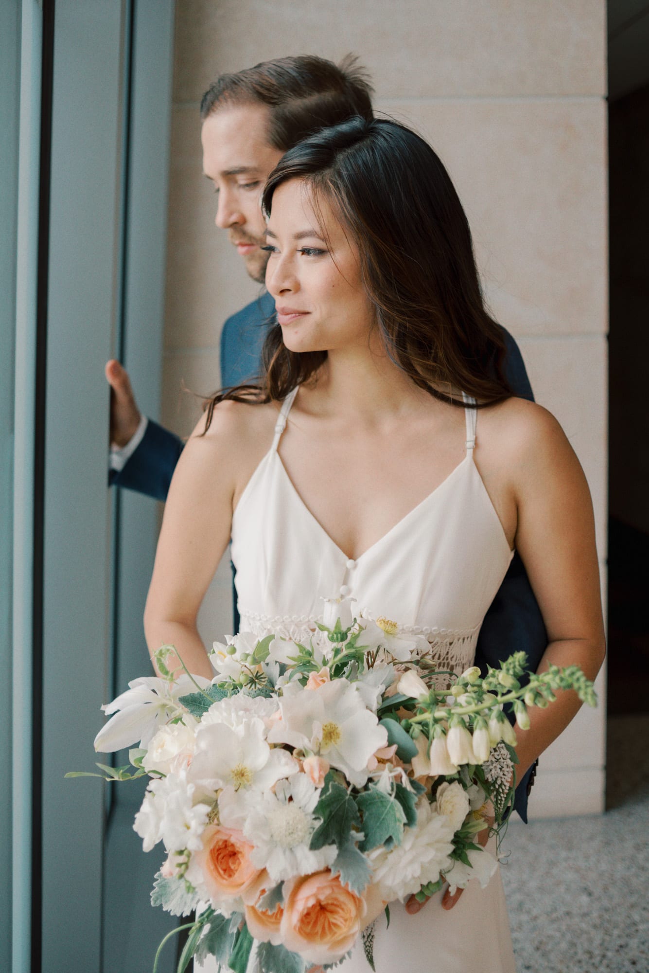 Newlyweds look out a window to the water while holding the white and pink bouquet