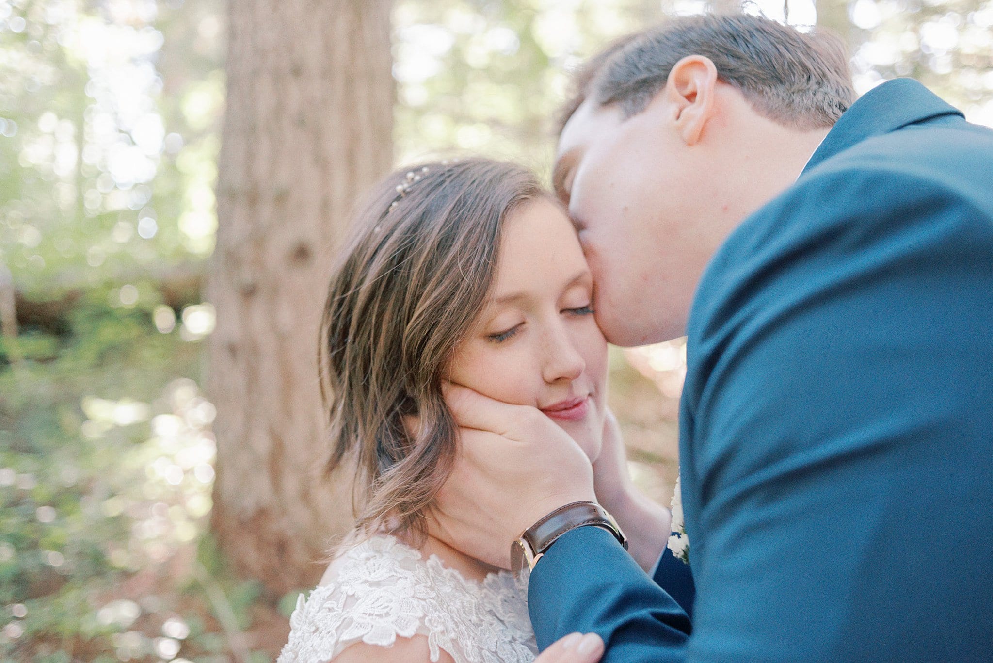 A groom in a blue suit kisses the temple of his bride with hands on her face at a Treehouse Point Wedding