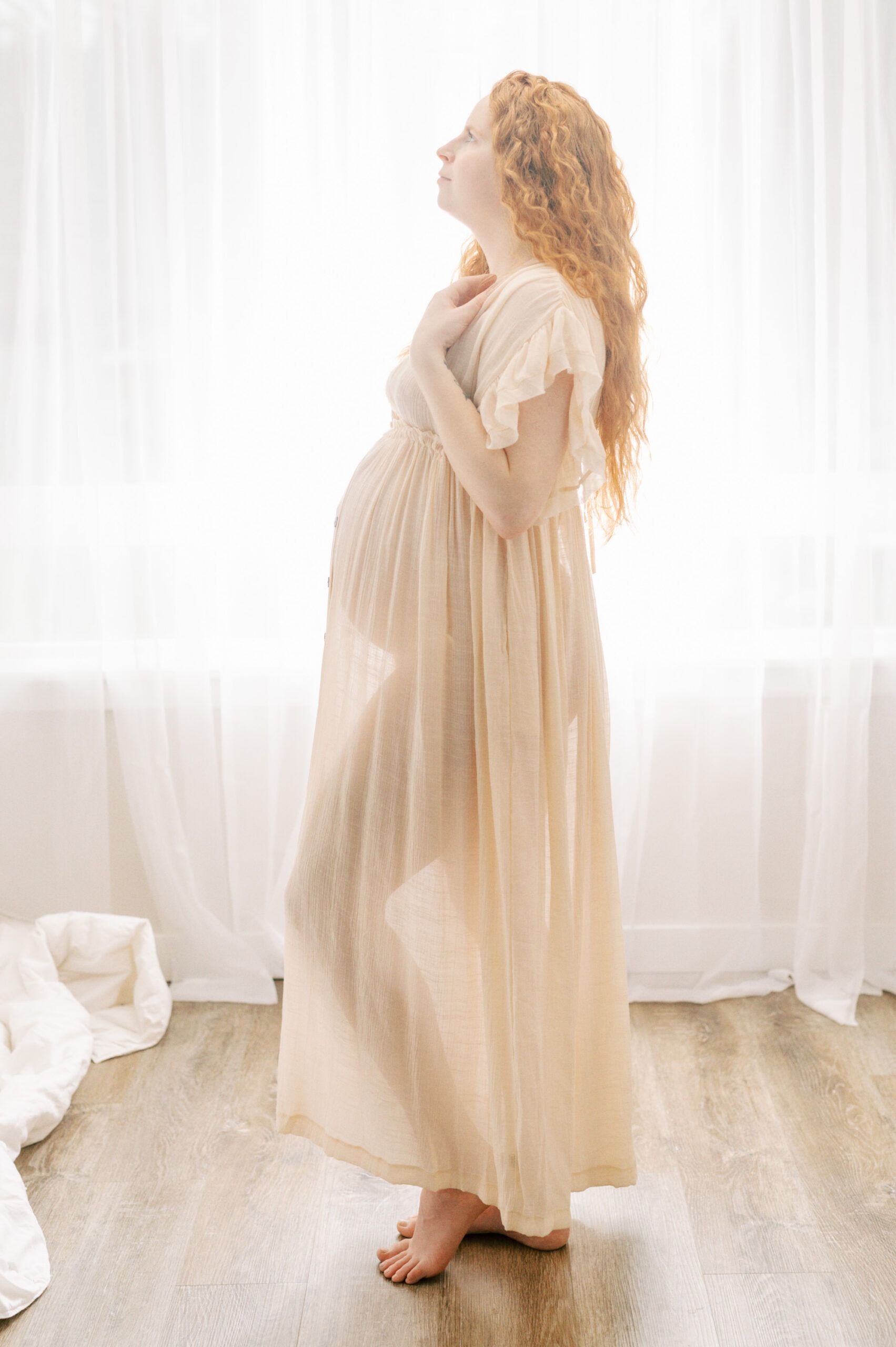 A mother to be stands in a studio window in a sheet cream dress