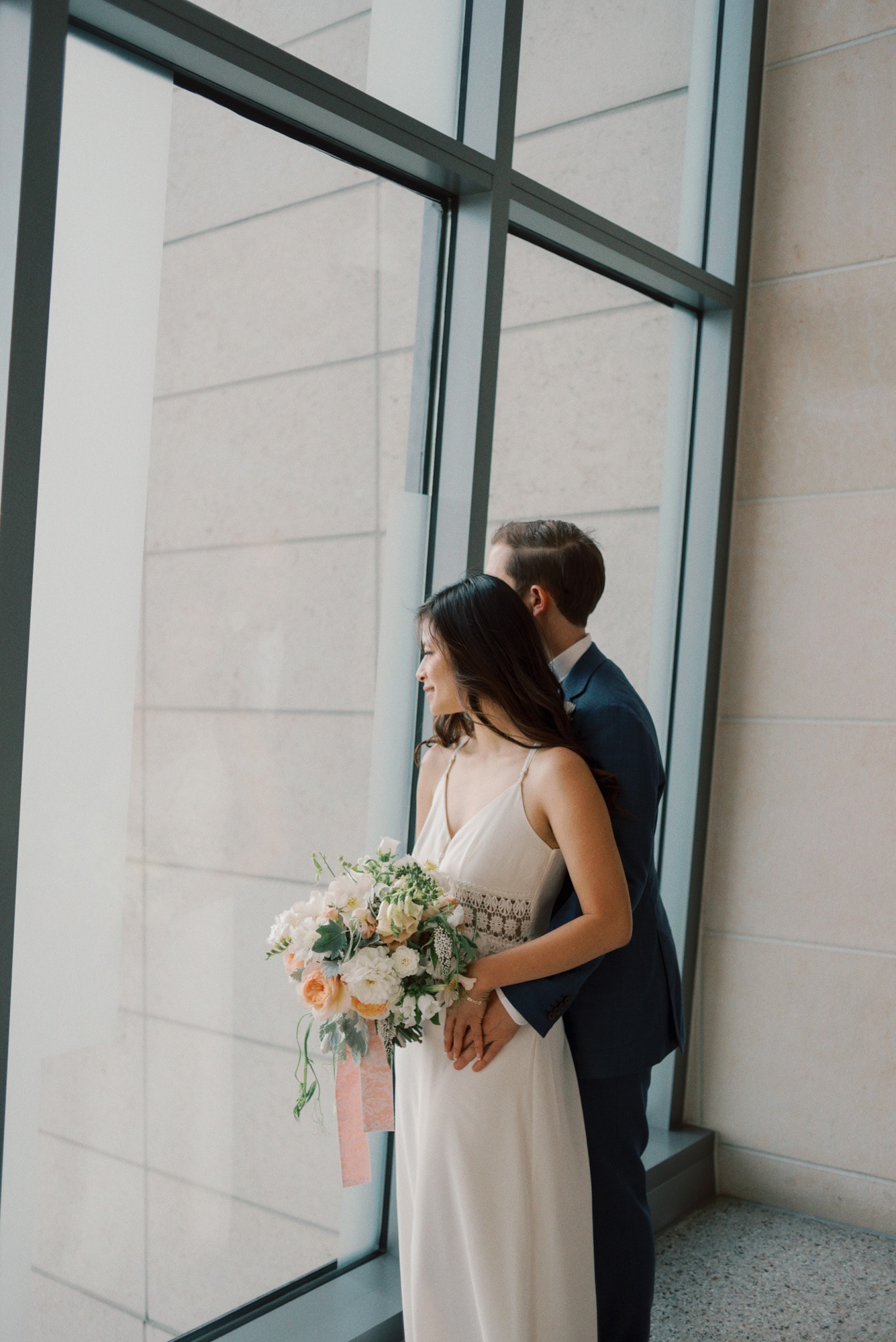 A bride and groom stand looking out a large window while holding each other and the colorful bouquet at The 101 Wedding Venue